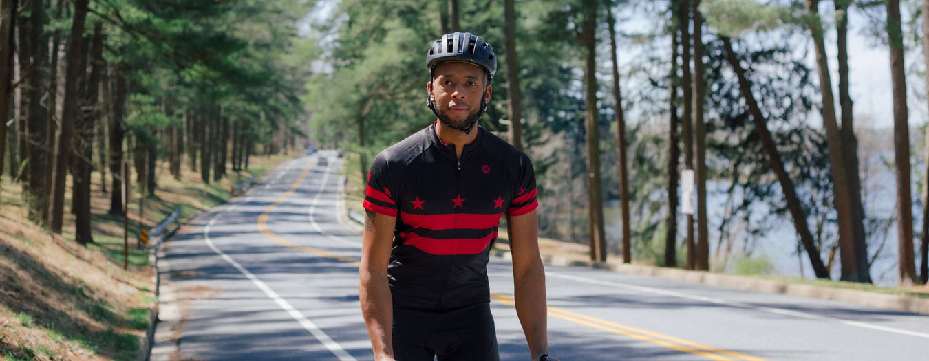 A cyclist wearing a black Washington DC flag cycling jersey pedals up a hill on a road surrounded by pine trees.